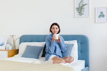 Obraz premium Dziewczyna w niebieskim swetrze we wnętrzu stylu Hygge z filiżanką w ręku siedzi na łóżku