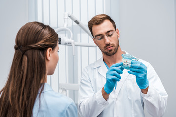 selective focus of bearded dentist in glasses holding tooth model near patient