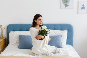 A girl in a white sweater sits on a bed holding a bouquet of white roses in her hand