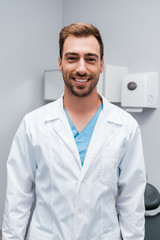 happy bearded doctor in white coat looking at camera while standing in clinic