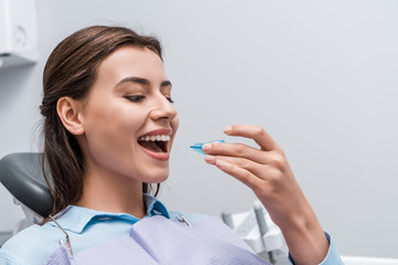 cheerful woman with opened mouth holding retainer in dental clinic - obrazy, fototapety, plakaty