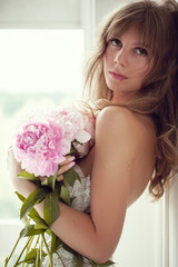 Beautiful blonde woman with sensual lips posing in lightfull white room among peonies flowers. - Image