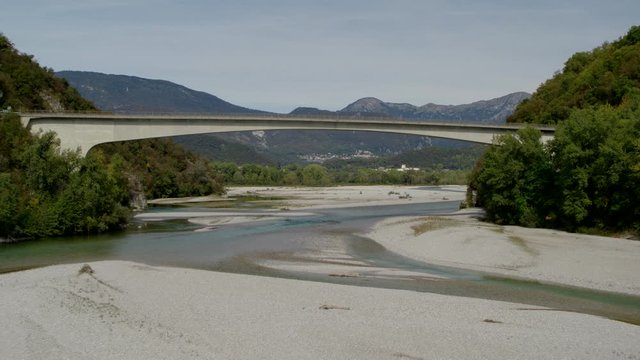 Drone flies over a small lake under a bridge with a scenic view of mountains in the background. Tagliamento, Italy.
