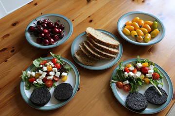 Two plates of healthy organic breakfast of black pudding with mixed leaf salad topped with yellow & red cherry tomatoes & feta cheese on a white plate.  Background of sliced bread, cherries & tomatoes