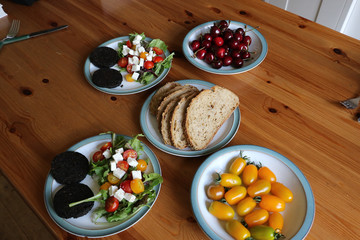 Two plates of healthy organic breakfast of black pudding with mixed leaf salad topped with yellow & red cherry tomatoes & feta cheese on a white plate.  Background of sliced bread, cherries & tomatoes