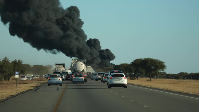 Traffic on highway comes to standstill as thick, black smoke and fireballs billow into the air from along the roadway. Cars and trucks in traffic jam, viewed from behind.