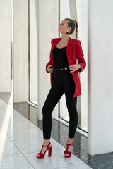 young european girl in red jacket, heels and black trousers posing near panoramic glazing business center in full growth. Classic female look.