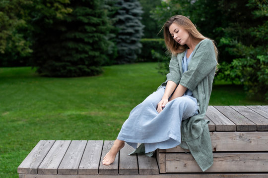 young European girl, natural long hair, beautiful face. blue long dress, gray jacket, sit on wooden platform in green park. Linen womens clothing. hipster lifestyle.