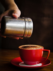 Hand sprinkles chocolate powder on drink in red cup.