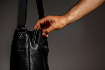 Hand of a thief pulling a purse from a black leather bag on a gray background