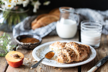 Fresh bread and croissant with milk and peach or apricot jam on a wooden table. Bouquet of daisies. rustic