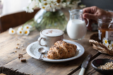 Breakfast cup of coffee and croissant with milk and peach or apricot jam on a wooden table. Bouquet of daisies. rustic