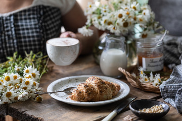 Breakfast cup of coffee and croissant with milk and peach or apricot jam on a wooden table. Bouquet of daisies. rustic
