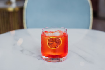 An orange aperitif cocktail in a rocks glass served on an ice cube with an orange slice in a bright colorful surrounding of a bar. Natural light. Marble table.