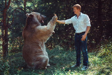 Red-haired man in a white shirt gives brown bear a high five in the forest. The theme of friendship...