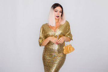 Charming young blonde haired woman wearing elegant gold evening dress and holding gold color clutch...