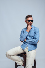 Confidence. Portrait of charming bearded man in casual clothes and sunglasses touching his chin while sitting on the chair against grey background