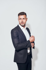 In his own style. Handsome bearded businessman in classic suit adjusting sleeves and looking away while standing against grey background