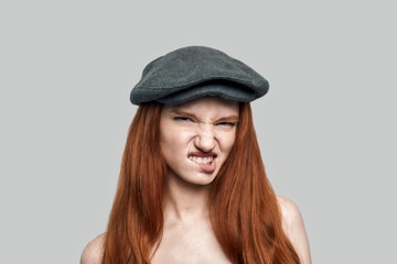 Rude girl. Portrait of young dissatisfied redhead woman in headgear biting a lip and looking at camera while standing against grey background
