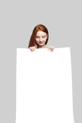 Pretty young redhead woman holding blank sign board and looking at it while standing against grey background