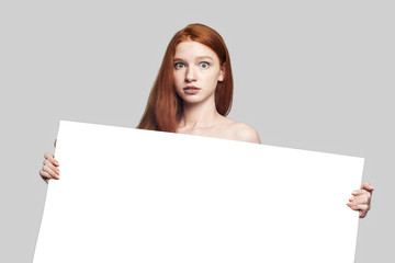 Fototapeta premium Your text here. Beautiful and young redhead woman holding empty blank board and looking at camera while standing against grey background