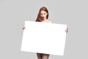 Shocked young redhead woman holding empty blank board and looking at it while standing against grey background