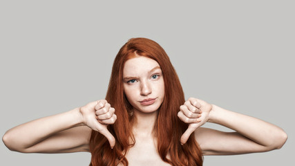 It is not cool. Studio shot of redhead girl showing thumbs down and making upset face while standing against grey background