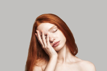 I am tired. Portrait of young frustrated teenage girl with long red hair keeping eyes closed and touching her face while standing against grey background