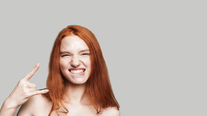 Rock star. Portrait of young and cute teenage girl with long red hair showing hand sign and looking at camera while standing against grey background