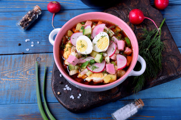 Russian cold soup Okroshka with vegetables, sausage, eggs and kvass in a ceramic bowl on the blue table