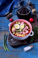 Russian cold soup Okroshka with vegetables, sausage, eggs and kvass in a ceramic bowl on the blue table
