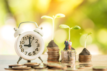 Miniature people: happy couple on stacked of coins with white clock next to, banking and saving concept.