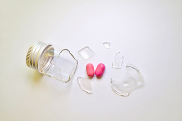 A broken glass bottle with a metal cap, two pink heart-shaped tablets fell out of it, on a white...