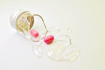 A broken small glass bottle with a metal cap, two pink tablets fell out of it, on a white background