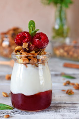 Layered breakfast with granola, yogurt and jam topped with raspberries in a glass jar on the white table