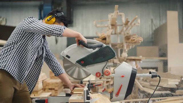 Guy in protective glasses and headphones is sawing timber with circulat saw in wood workshop making handmade furniture. People, woodwork and equipment concept.