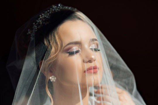 Beautiful bride in veil. Close Up shot of elegant bride in luxury white dress posing under veil close up. Bride portrait wedding makeup and hairstyle with diamond crown, fashion bride.