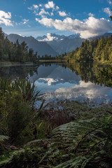 Fototapeta na wymiar A portrait view of the incredibly beautiful Lake Matheson, New Zealand with the reflection of the stunning Southern Alps and the majestic Mt Cook in the still waters, foliage in the foreground