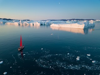 Little red sailboat cruising among floating icebergs in Disko Bay glacier during midnight sun season of polar summer. Ilulissat, Greenland. Arctic nature ice landscape in Unesco World Heritage Site.