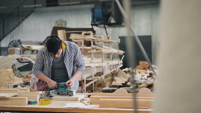 Zoom-out of male carpenter in safety headphones polishing timber with belt sander working in factory alone busy with work. People, business and job concept.