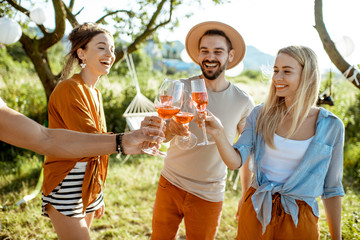 Fototapeta premium Young and cheerful friends having fun, clinking with wine glasses in the beautifully decorated backyard during a festive meeting or party on a sunny summer evening