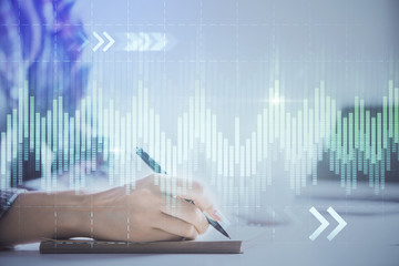 A woman hands writing information about stock market in notepad. Forex chart holograms in front. Concept of research. Double exposure