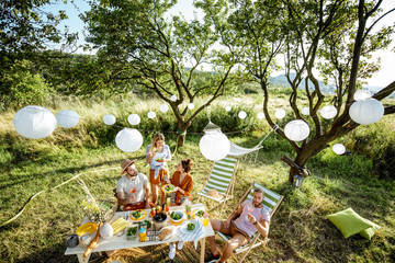 Group of a young friends having festive lunch in the beautifully decorated garden on a summer afternoon, wide view from above