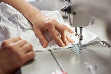 Close up side top view of cropped female hands sewing white fabric on professional manufacturing machine at seamstress workplace. Tailor hands holding textile for dress production. Blurred background