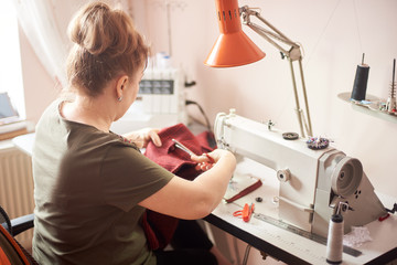 Back view of tailor laboring at working place in atelier. Female hands using scissors for cutting off cloth piece in sewing process. Sewing machine, ruler, scissors, table lamp, threads on background.