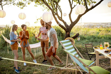 Young and happy friends having fun, dancing together with wine during a picnic in the beautiful garden on a sunset