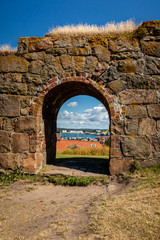Fototapeta premium Summer view through an old medieval stone wall arched gate at Varberg Fortress in Sweden with the city and harbor in the background.