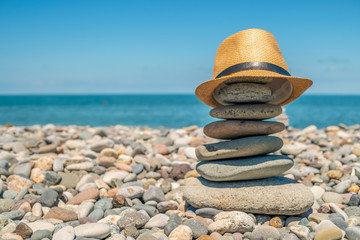 Fototapeta na wymiar A cairn on a pebble beach against the sea on a sunny day, a straw hat is put on the cairn. Travel and Vacation Concept