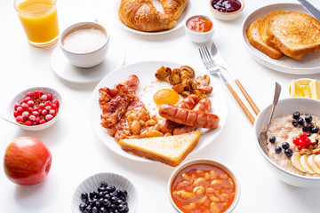 Top view flatlay with varieties of fresh breakfast: fried eggs with bacon and sausages, oatmeal with berries, fried toasts with jam and butter. White background.