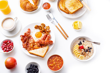Top view flatlay with varieties of fresh breakfast: fried eggs with bacon and sausages, oatmeal with berries, fried toasts with jam and butter. White background.
